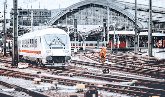 Ein weißer ICE fährt an einem Hauptbahnhof los. Im Hintergrund arbeiten Gleisarbeiter an den Gleisen